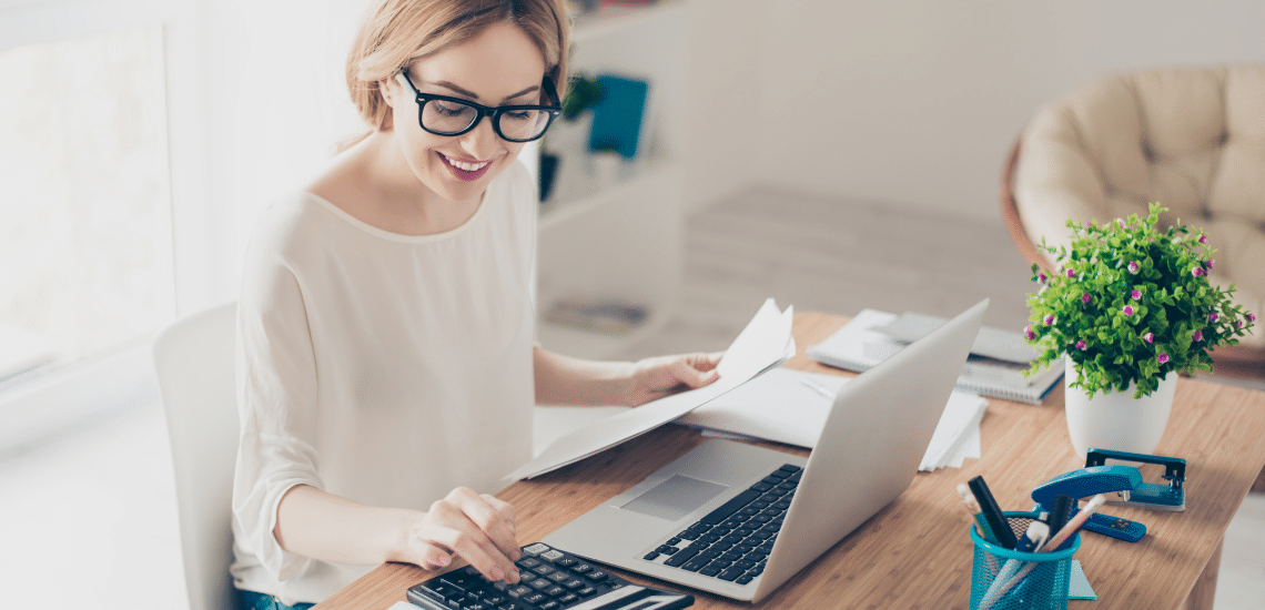 A woman with glasses sitting at a desk with a calculator and a laptop
