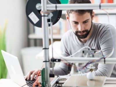 A man using a laptop and operating a 3D printer in a workshop